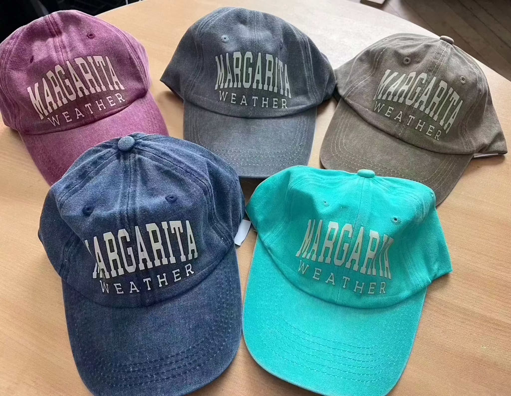 Five Margarita Weather cotton baseball caps in various colors displayed on a wooden surface