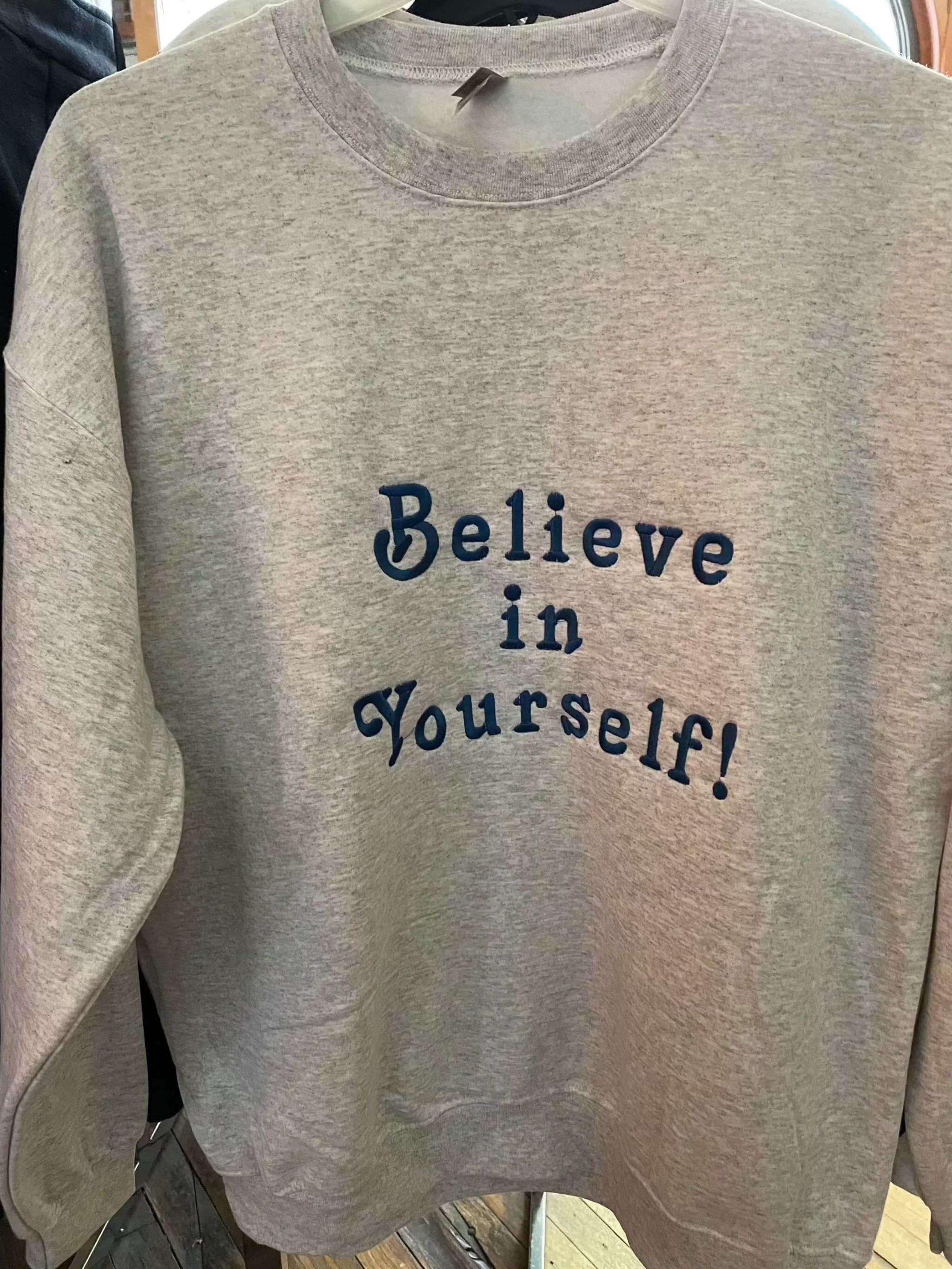 Grey crewneck sweatshirt with "Believe in Yourself!" text in blue lettering
