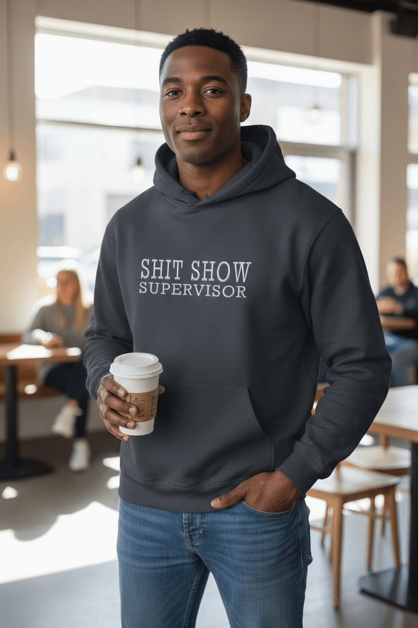 Man wearing black SHIT SHOW SUPERVISOR hoodie holding coffee cup indoors