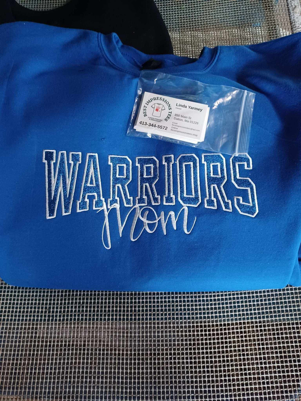 Blue Warriors Mom sweatshirt with white lettering and custom embroidery, shown folded with business card on top